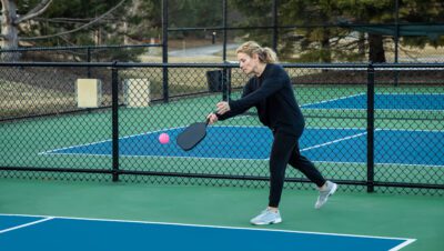 A female pickleball player prepares to serve a bright pink ball on a blue and green court in early spring.